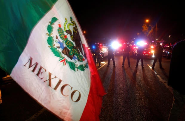 Police in riot gear form line to break up group of protesters, one with a Mexican flag, outside Trump's rally in Costa Mesa
