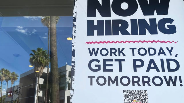 Sign reading "Now hiring. Work today, get paid tomorrow!" with palm tree reflection in the background 
